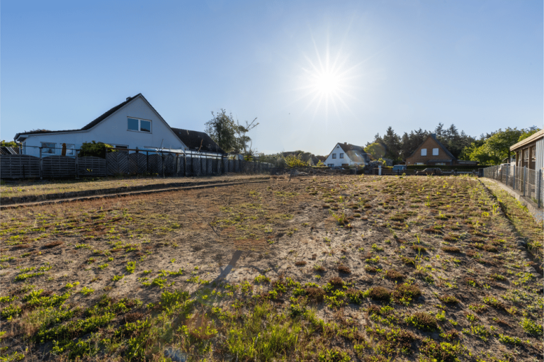 Blick auf einen leeren Grundstück mit Häusern im Hintergrund und strahlender Sonne.